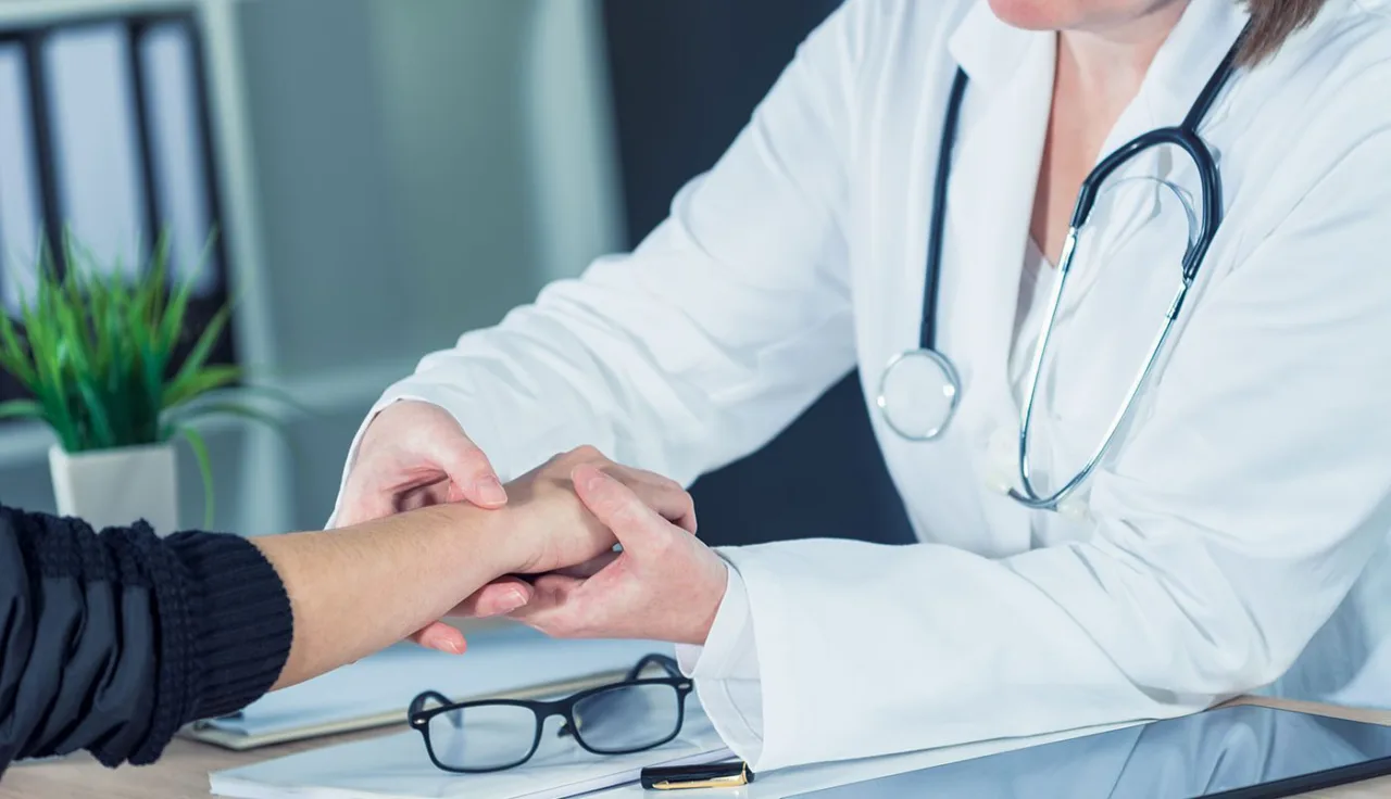 Doctor in a white coat examining a patient's hand across a consulting room desk