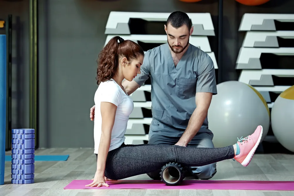Trainer guiding a woman through a foam-roller exercise on a gym floor