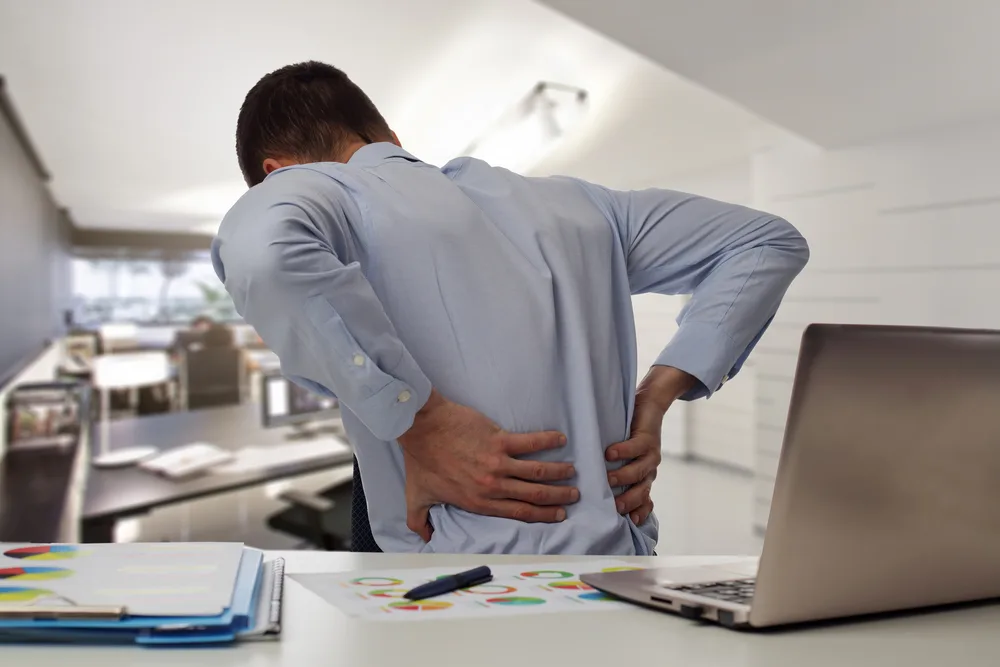 Man at an office desk holding his lower back beside a laptop