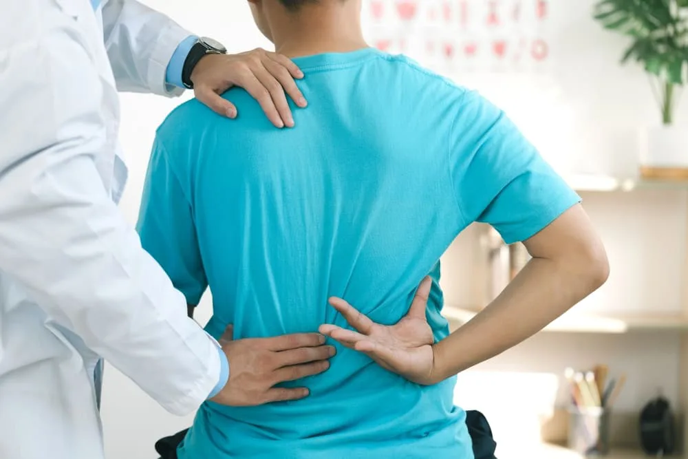 Person in a turquoise shirt holding their lower back while a clinician stands behind