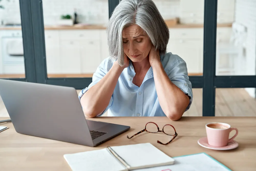 Woman at a laptop in a kitchen rubbing the back of her neck