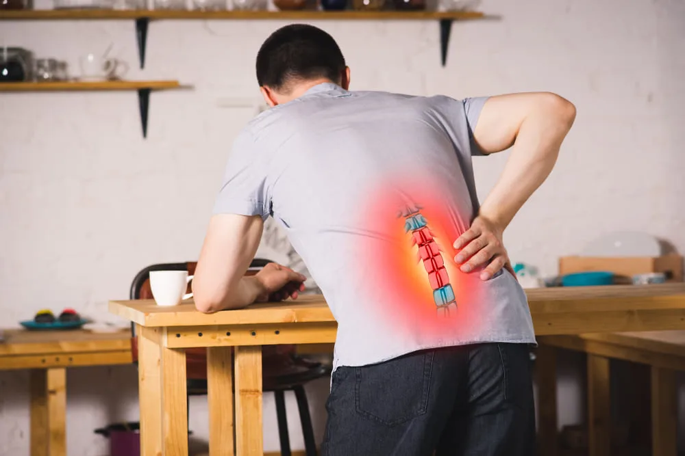 Man at a workbench holding his lower back, with a glowing overlay of the spine