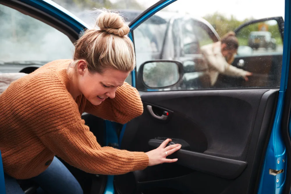 Woman in an orange sweater reaching for her back beside an open car door