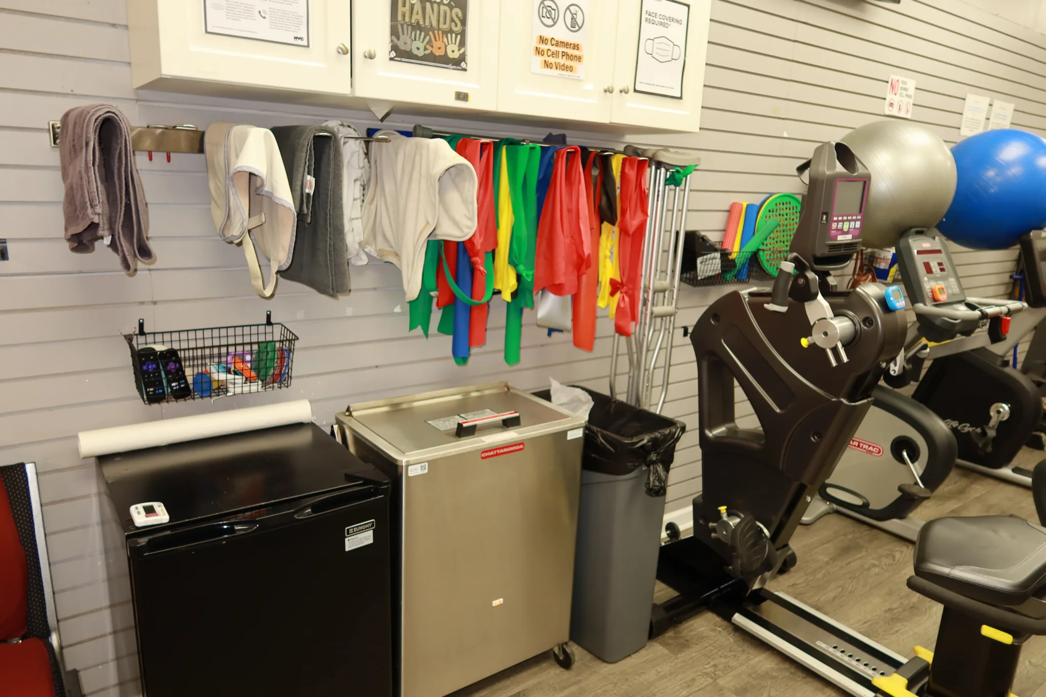 Slatwall of resistance bands and folded towels above a supply cart, beside an upper body ergometer