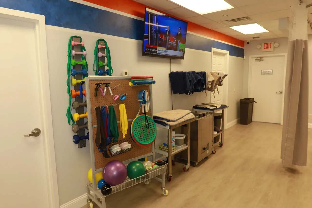 Pegboard of resistance bands and hand weights beside a supply cart and a wall-mounted screen