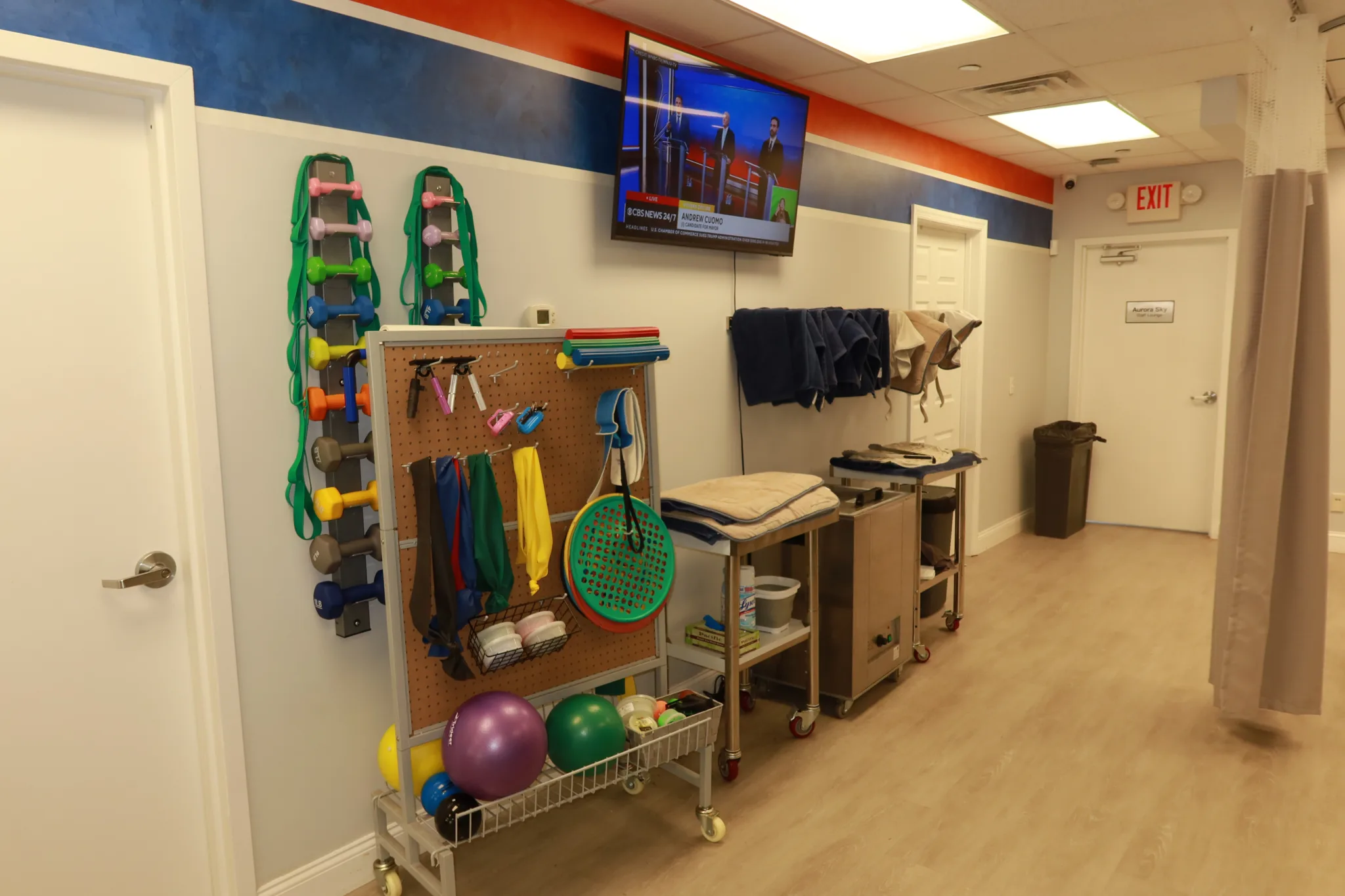 Pegboard of resistance bands and hand weights beside a supply cart and a wall-mounted screen