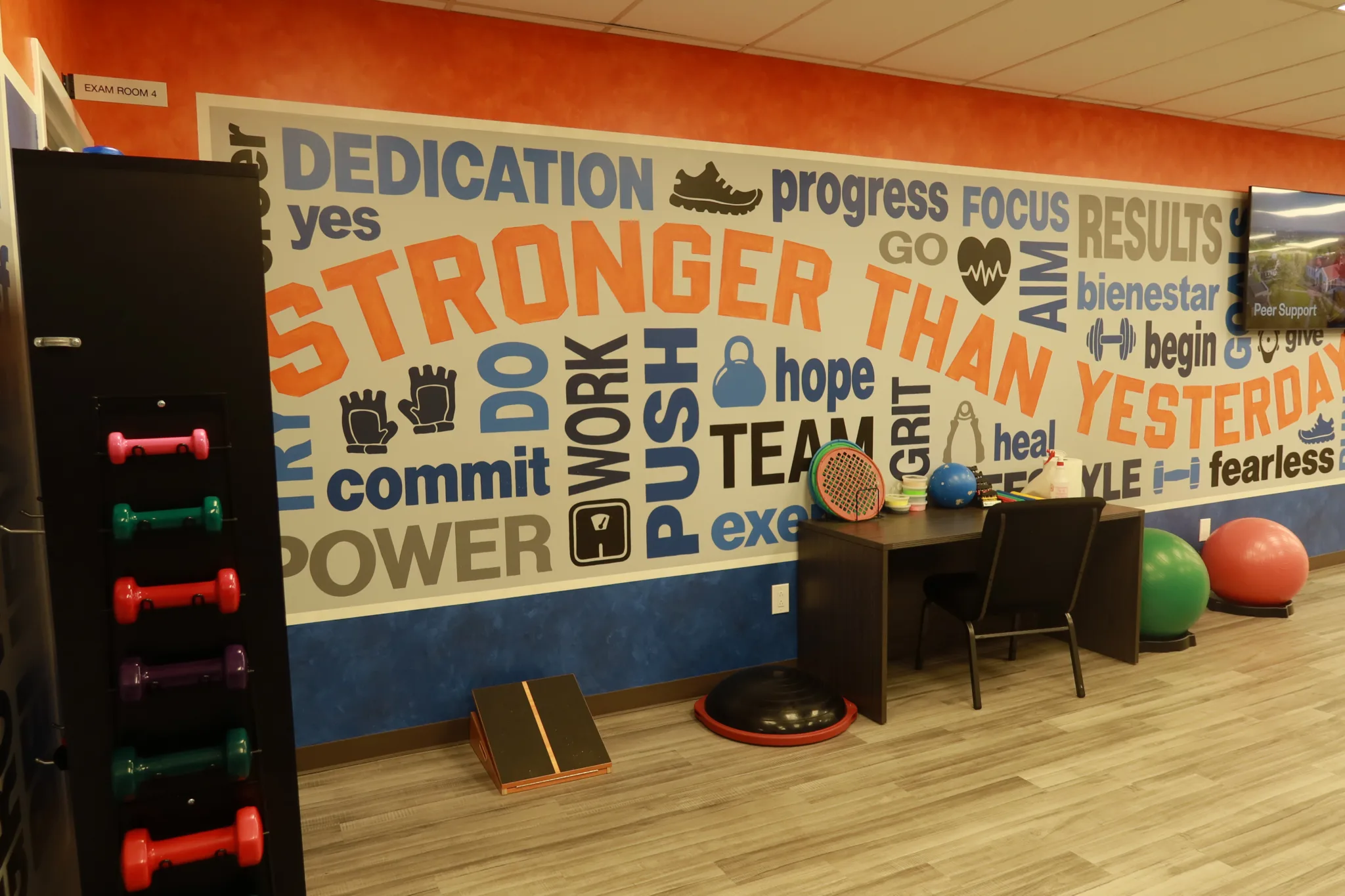 Wall mural reading Stronger Than Yesterday above a dumbbell rack, a work table and exercise balls