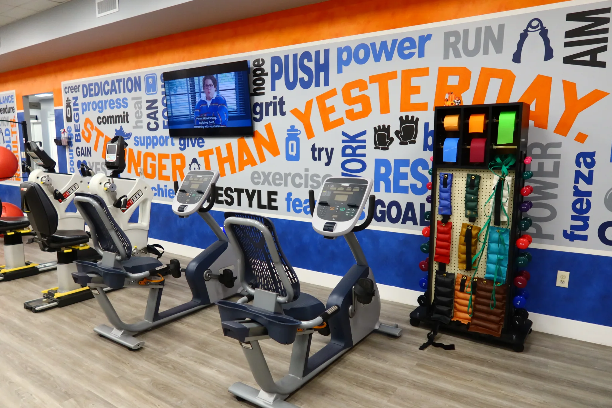 Exercise bikes in front of a Stronger Than Yesterday mural, beside a rack of resistance bands and weights