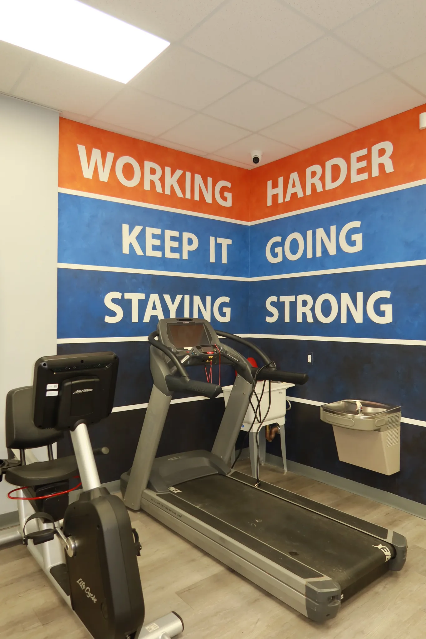 Treadmill in front of a mural reading Working Harder, Keep It Going, Staying Strong