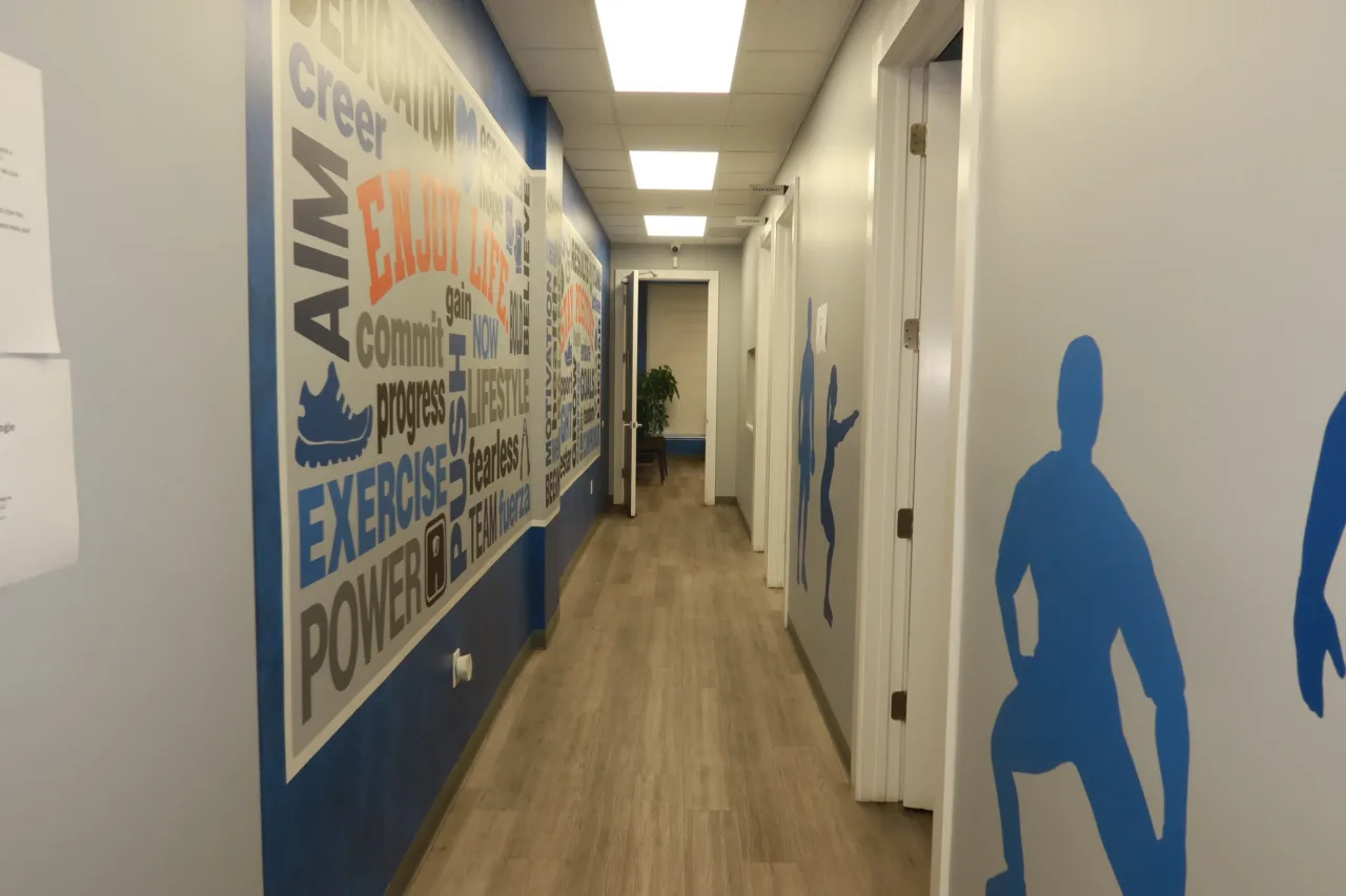 Hallway lined with blue athlete silhouettes and a bilingual word mural, leading to the treatment rooms