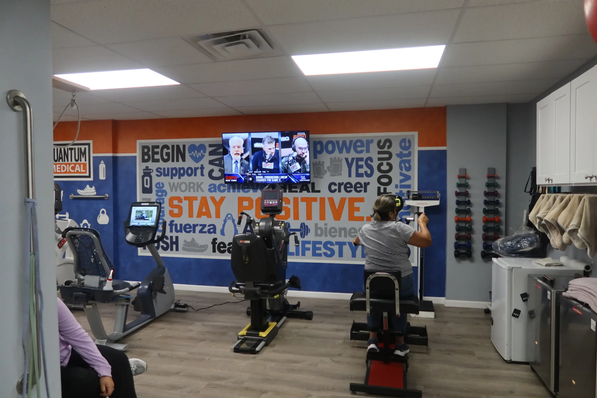 Cardio machines in front of a bilingual Stay Positive word mural and a wall-mounted screen, beside two racks of hand weights