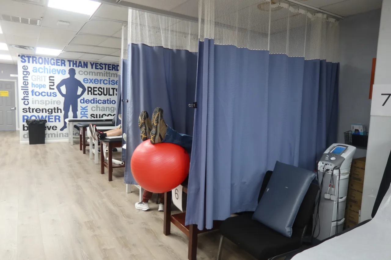 Row of curtained treatment bays, one with a patient's legs up on a red exercise ball, and a Stronger Than Yesterday mural beyond