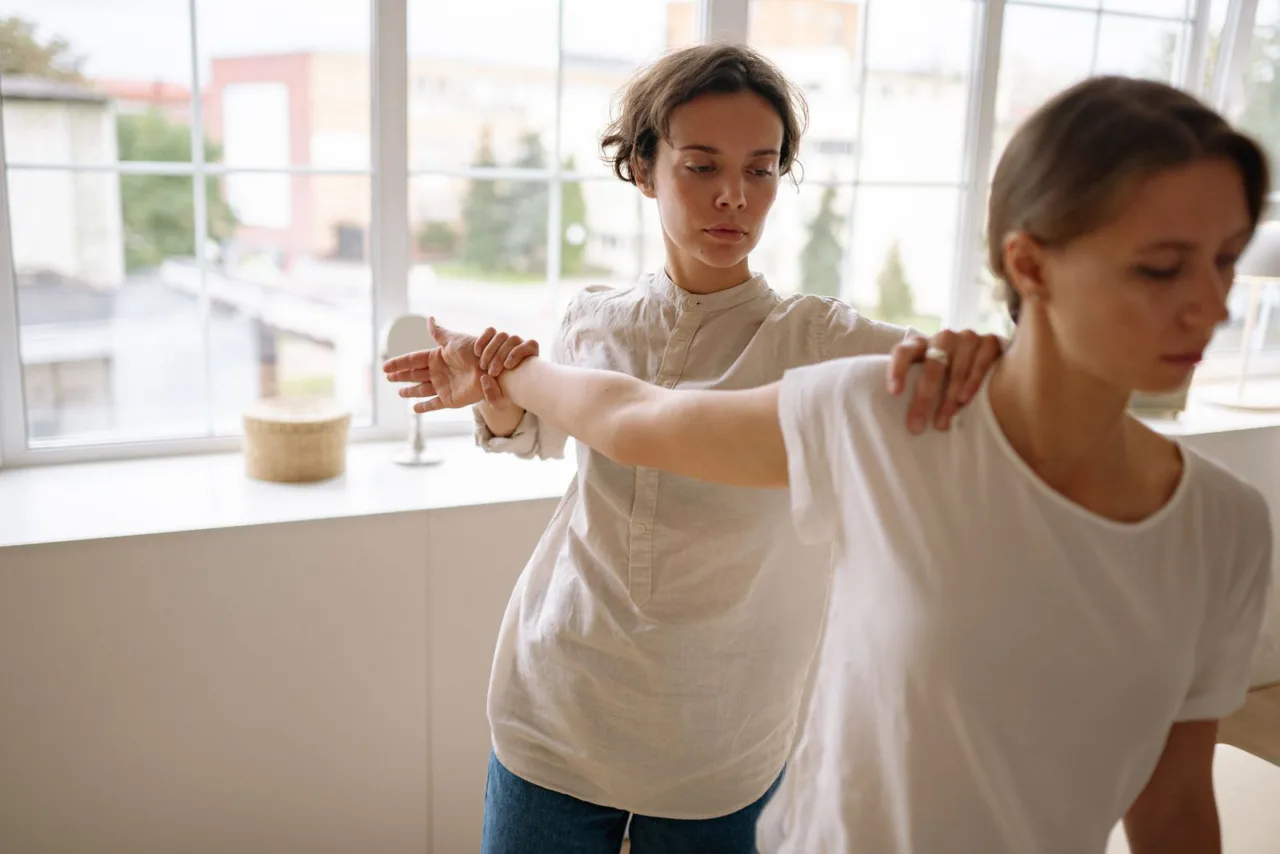 A therapist guides a patient's arms through a shoulder exercise beside a window.